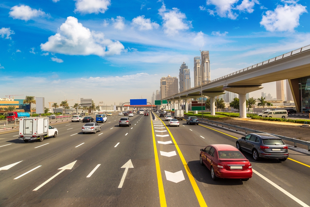 Traffic on a multi-lane highway in Dubai, with an elevated road and modern city buildings in the background under a blue sky.