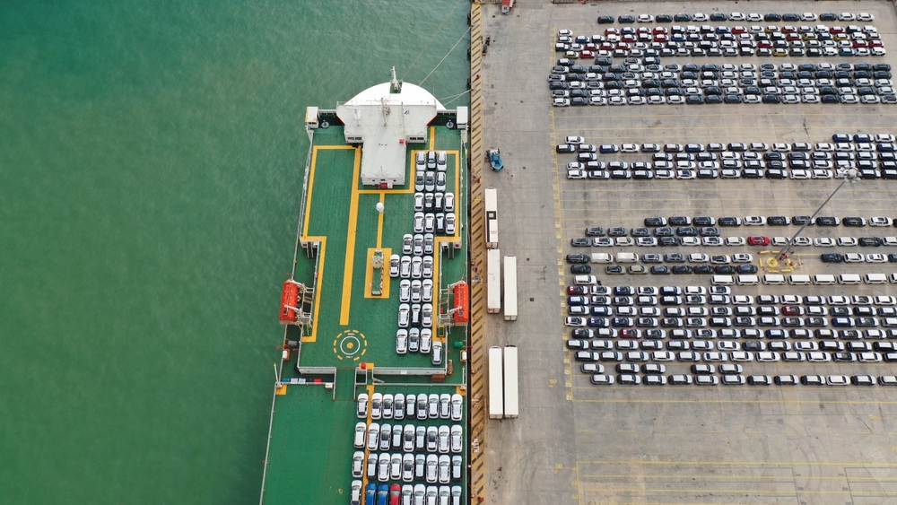 Aerial view of a car ferry docked beside a large vehicle parking area at a port, with rows of cars lined up near the water.