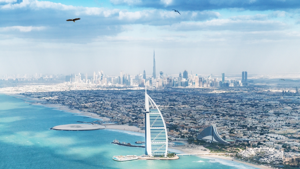 Aerial view of Dubai’s coastline featuring the Burj Al Arab in the foreground, with the city skyline and Burj Khalifa in the distance.